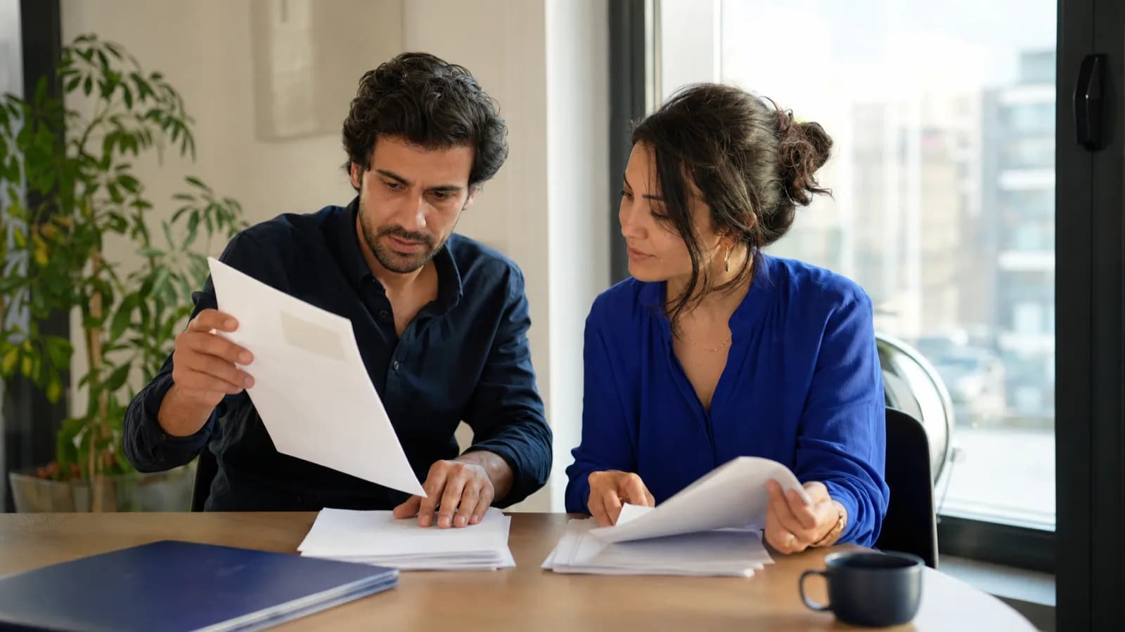 İki kişi ofis masasında basılı sayfa yığınını ikiye ayırıyor, biri elindeki sayfayı diğerine gösteriyor