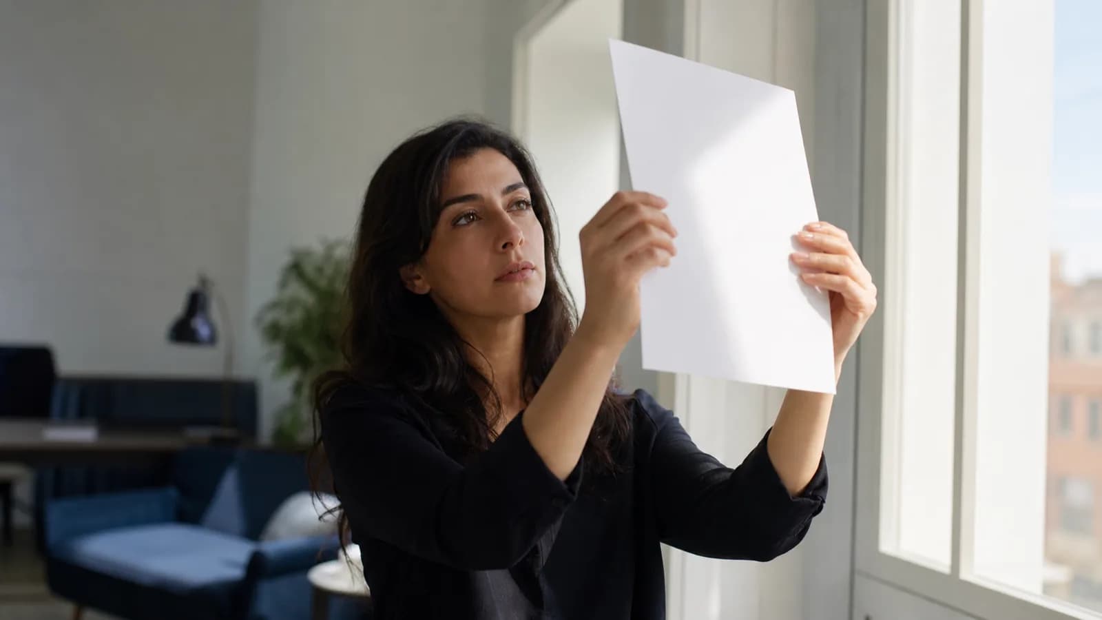A woman standing by a window, tilting the single page in her hand towards the light to study it.